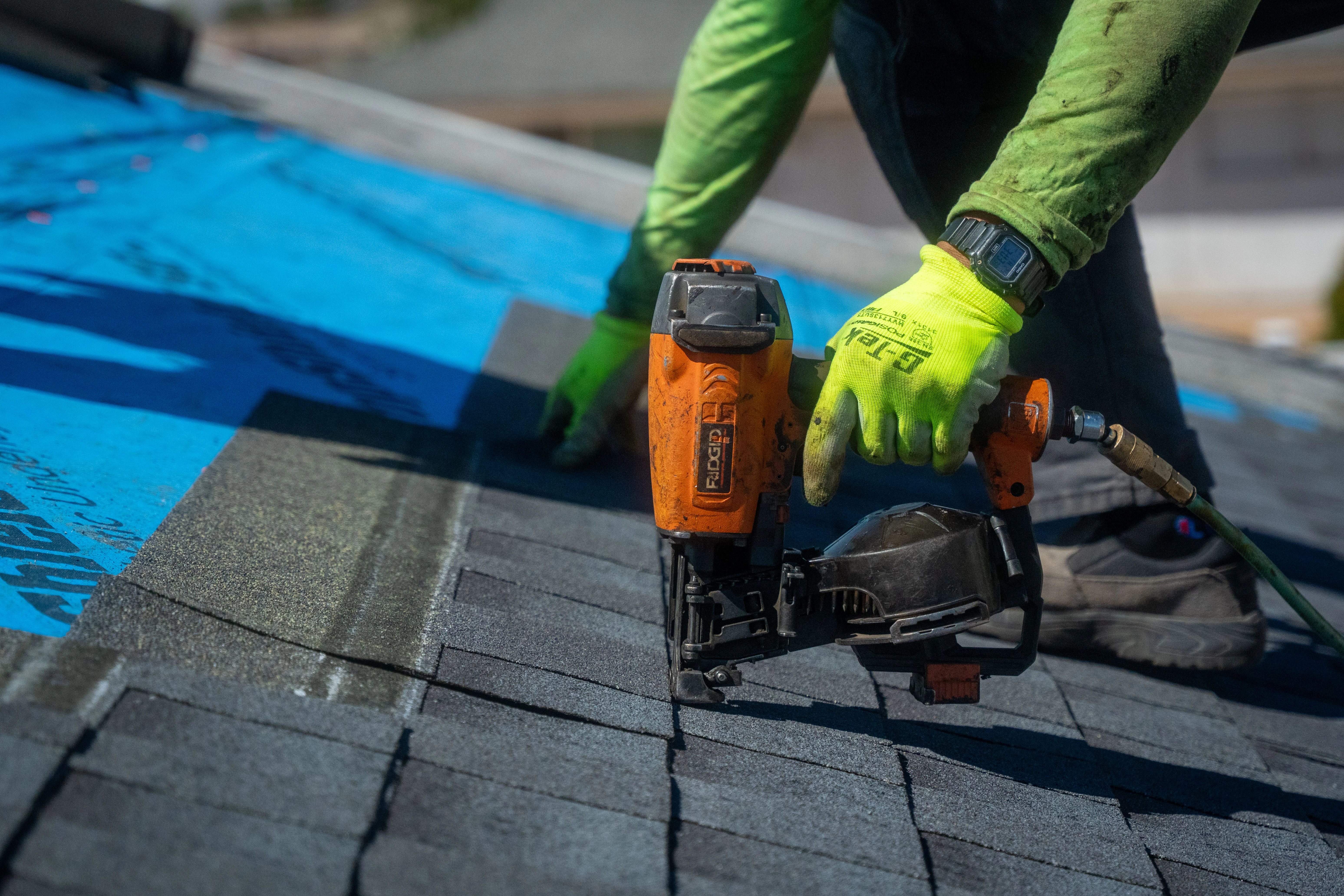 Roofing contractor installing asphalt shingles using a pneumatic nail gun on a residential roof during a roof replacement project